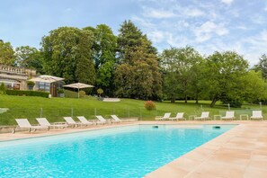 Una piscina con sillones de descanso, un área de estar techada y un edificio con jardín al fondo.