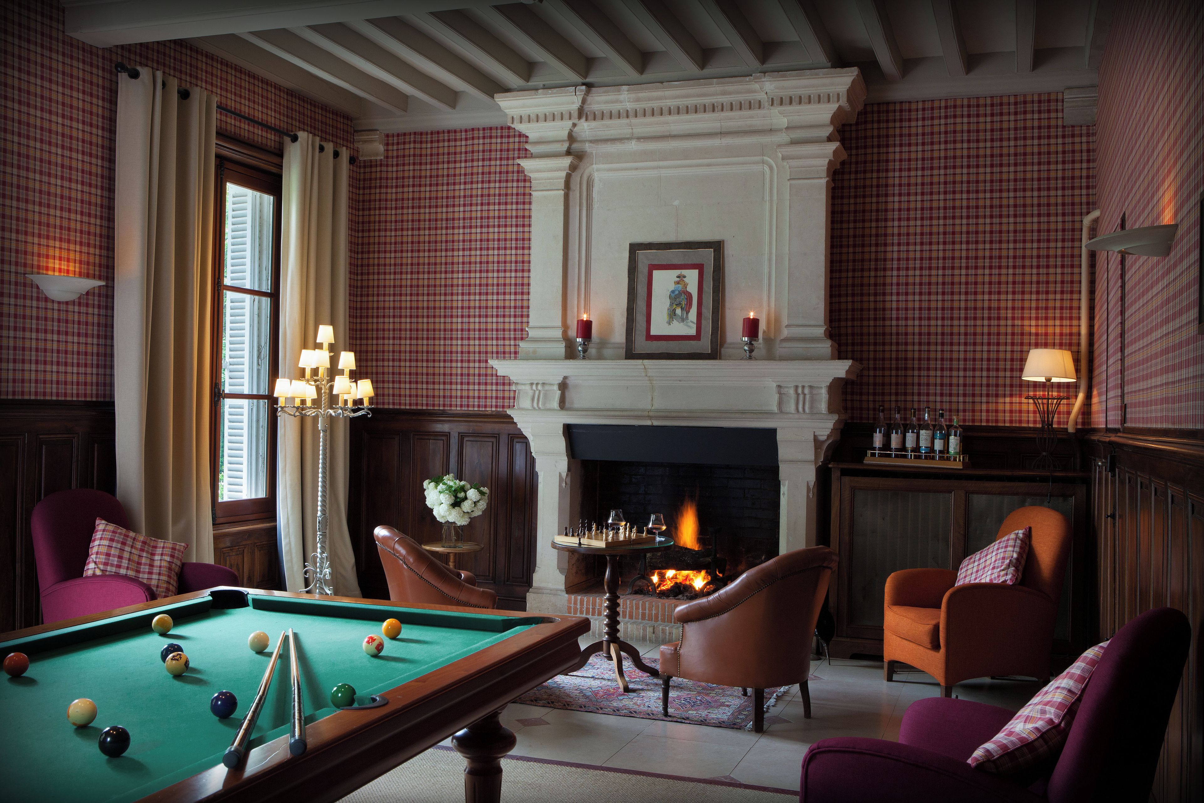 Une table de billard dans une pièce avec une cheminée, deux fauteuils et une table d’appoint sur laquelle est posé un vase de fleurs.