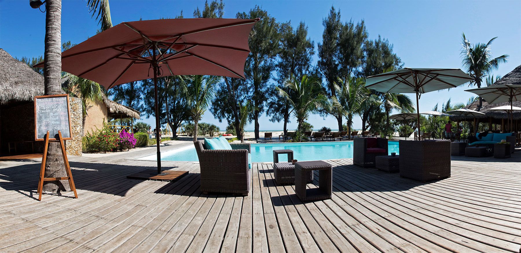 Een houten terras met lounge stoelen en parasols, een zwembad en een gebouw met rieten dak.
