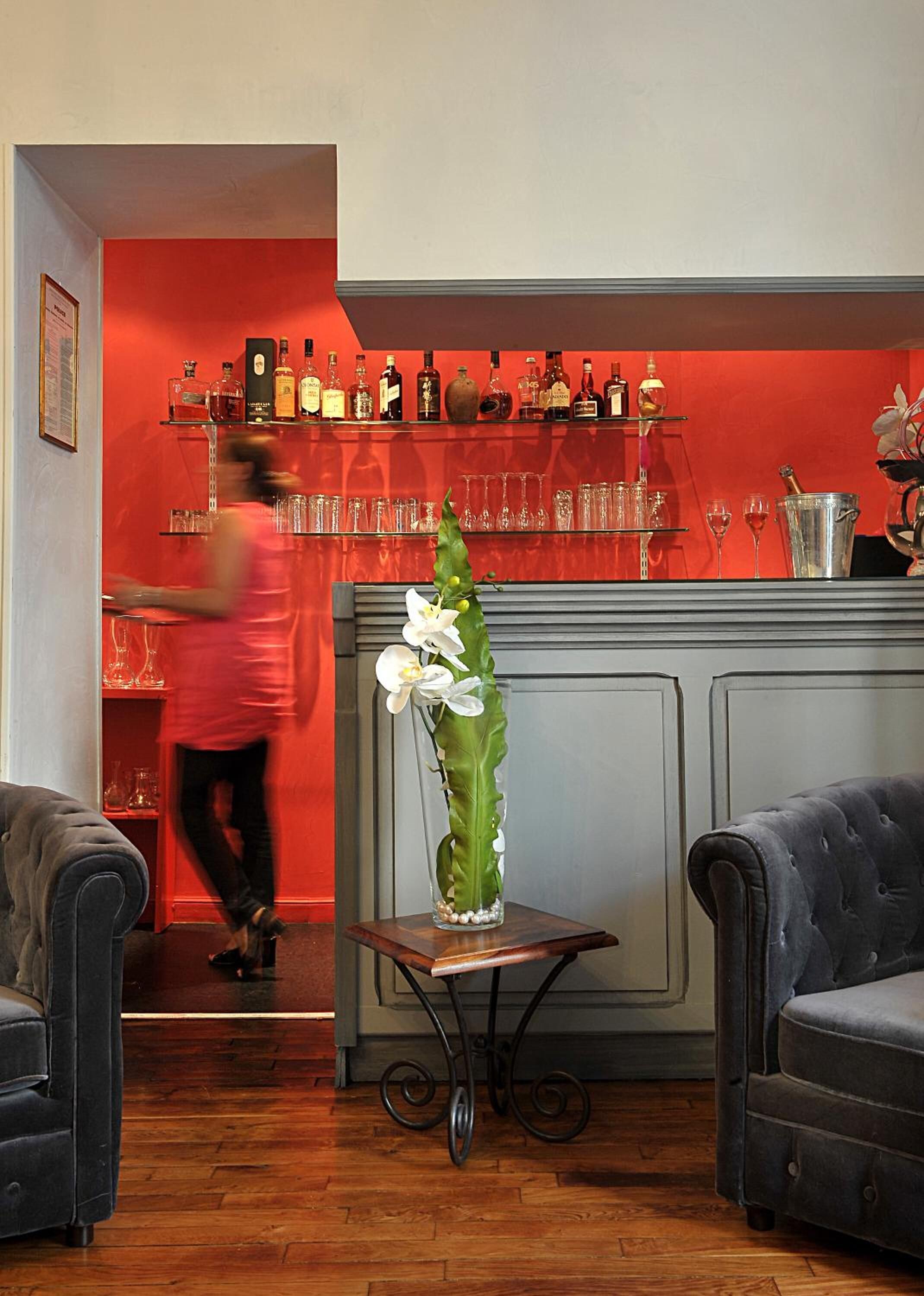 A bar area with a red wall, shelves of liquor bottles, and glasses. There is a table with a vase of flowers in the foreground.