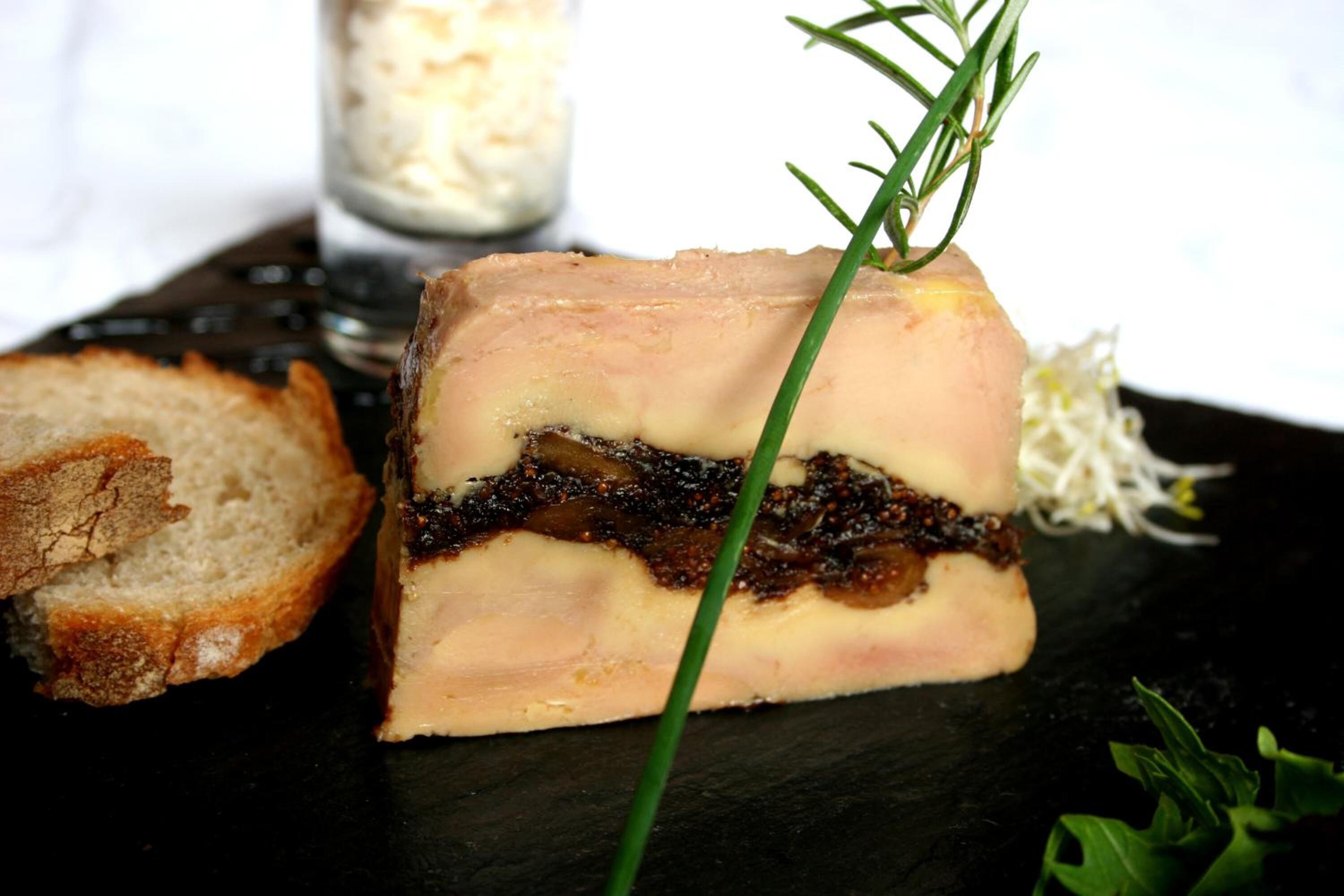 A block of pâté with a sprig of rosemary on top, accompanied by a slice of bread and some greens on a dark slate plate.