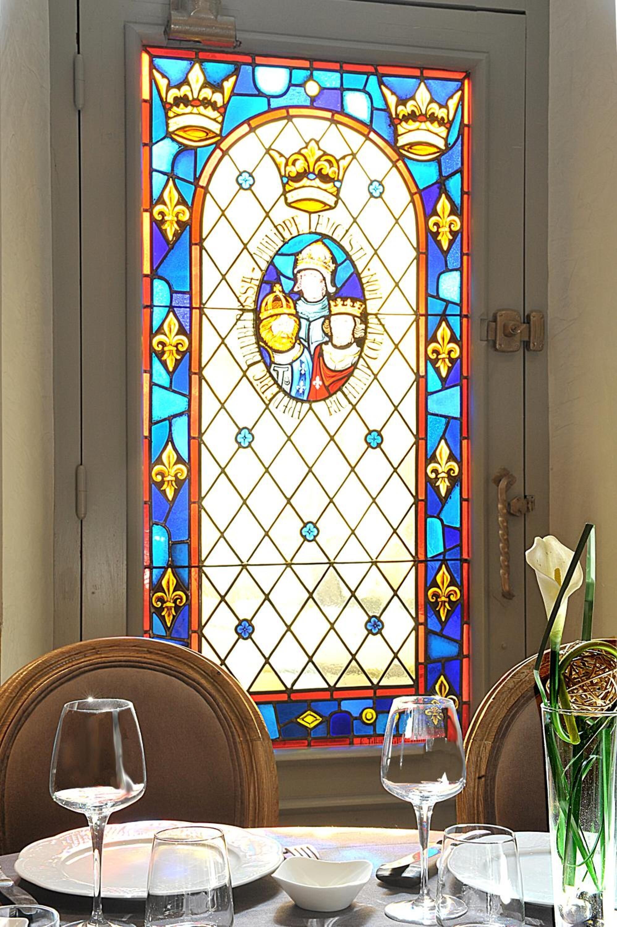 Un coin repas agrémenté d’une fenêtre à vitraux représentant une figure religieuse, d’une table dressée pour deux personnes et d’un vase rempli de fleurs.