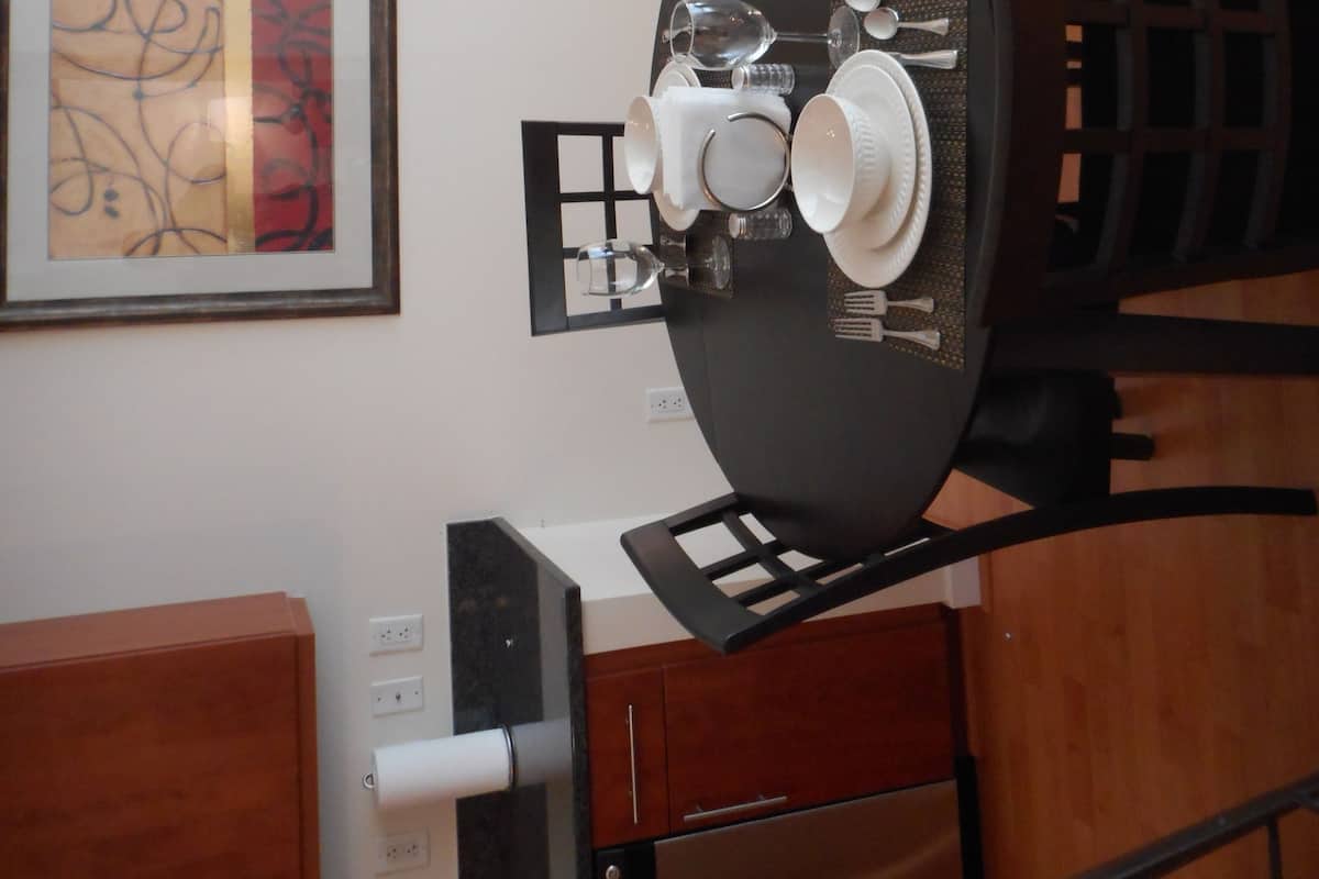 A dining area with a round table, chairs, and a light fixture. There is a framed artwork on the wall and a wooden cabinet below the table.