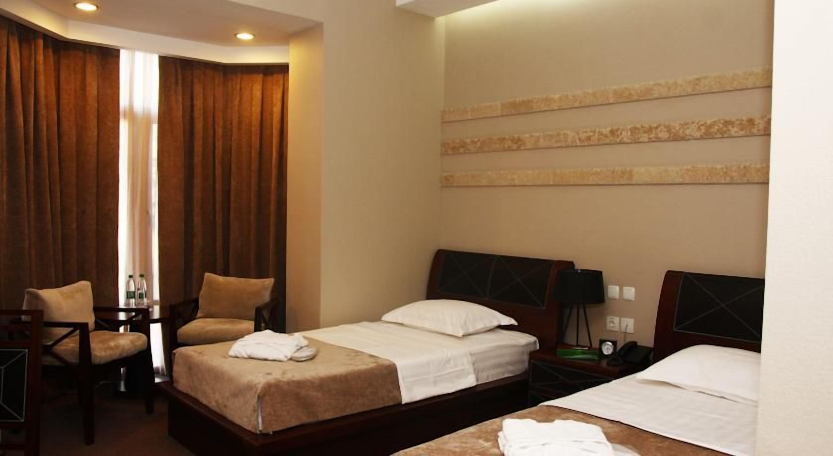 Quarto de hotel com cama, duas cadeiras, escrivaninha, mesa de cabeceira e luminária na parede.