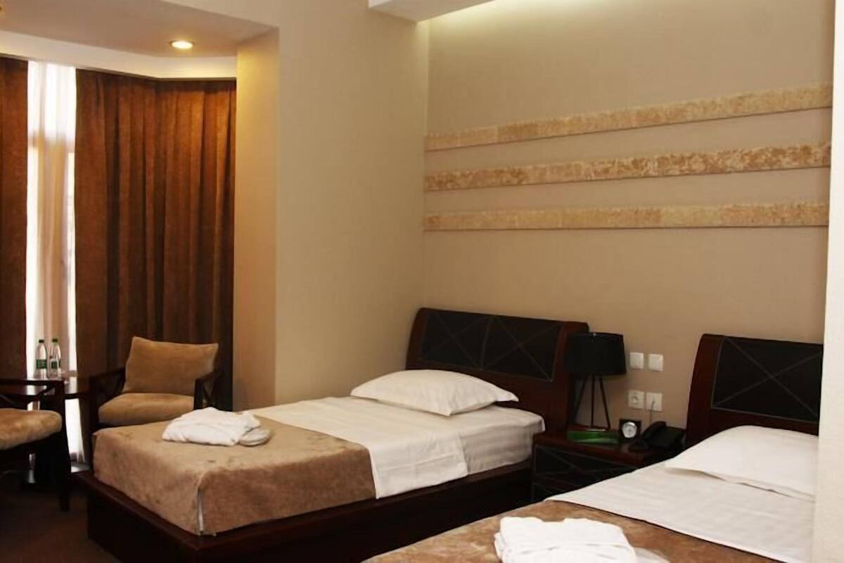 A hotel room with a bed, two chairs, a desk, a nightstand, and a wall-mounted lamp.