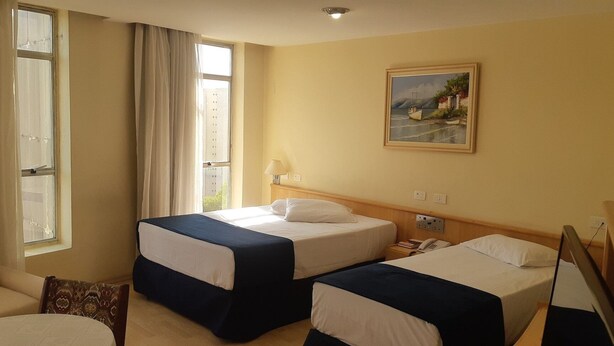 A hotel room with two beds, a window with curtains, a painting on the wall, and a bedside table with a lamp.