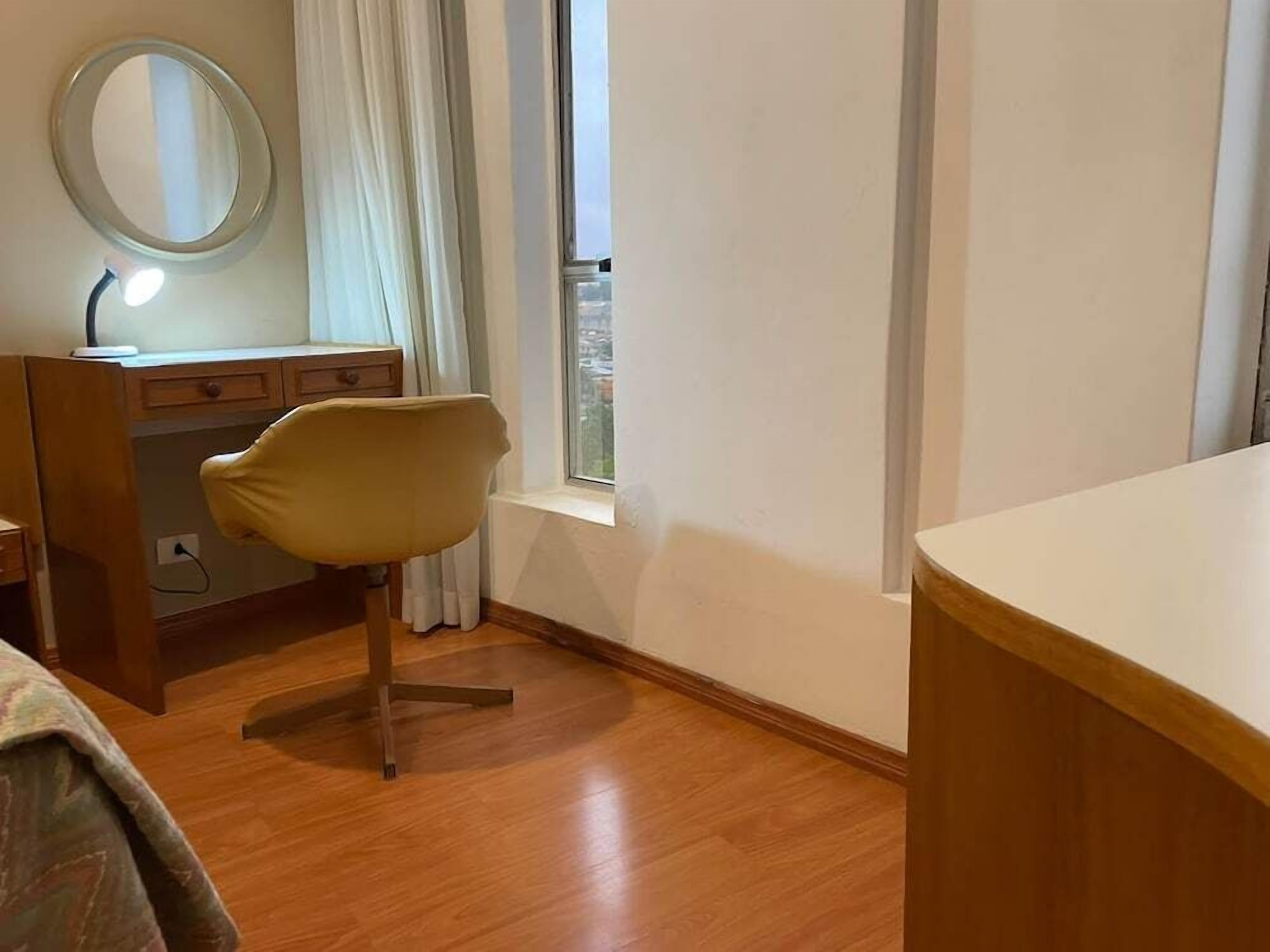 A bedroom with a wooden floor, a desk with a lamp, a chair, and a window with curtains.