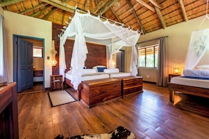 A room with a wooden floor, a bed with a canopy, a nightstand, a door, and a window. - Hakusembe River Lodge (Rundu)