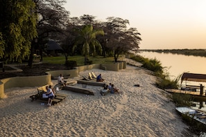 A sandy beach with lounge chairs and a boat, surrounded by trees and a calm river. - (Rundu)
