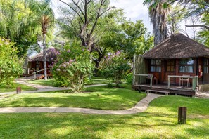 A bungalow with a thatched roof, surrounded by lush greenery and flowering plants. - Hakusembe River Lodge (Rundu)