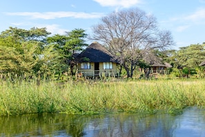 A thatched-roof building with a visible window and door, surrounded by tall grass and trees, near a body of water. - (Rundu)