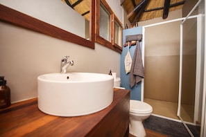 A bathroom with a wooden vanity, a white sink, a mirror, a towel rack, and a shower area. - Hakusembe River Lodge (Rundu)
