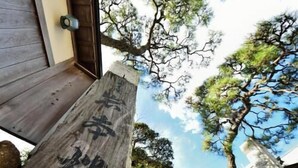 Un letrero de madera con caracteres japoneses, una puerta de madera y una vista de árboles y un edificio.