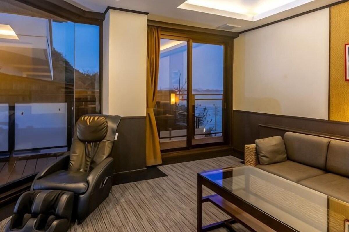 Habitación de hotel con sillón de masajes, sofá, mesa de centro de vidrio y una ventana grande con vistas.