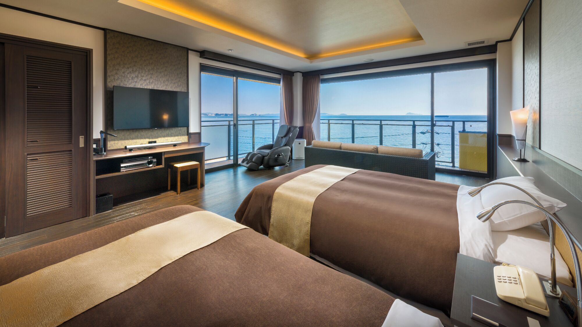 A hotel room with a large bed, a TV, a massage chair, and a view of the sea.