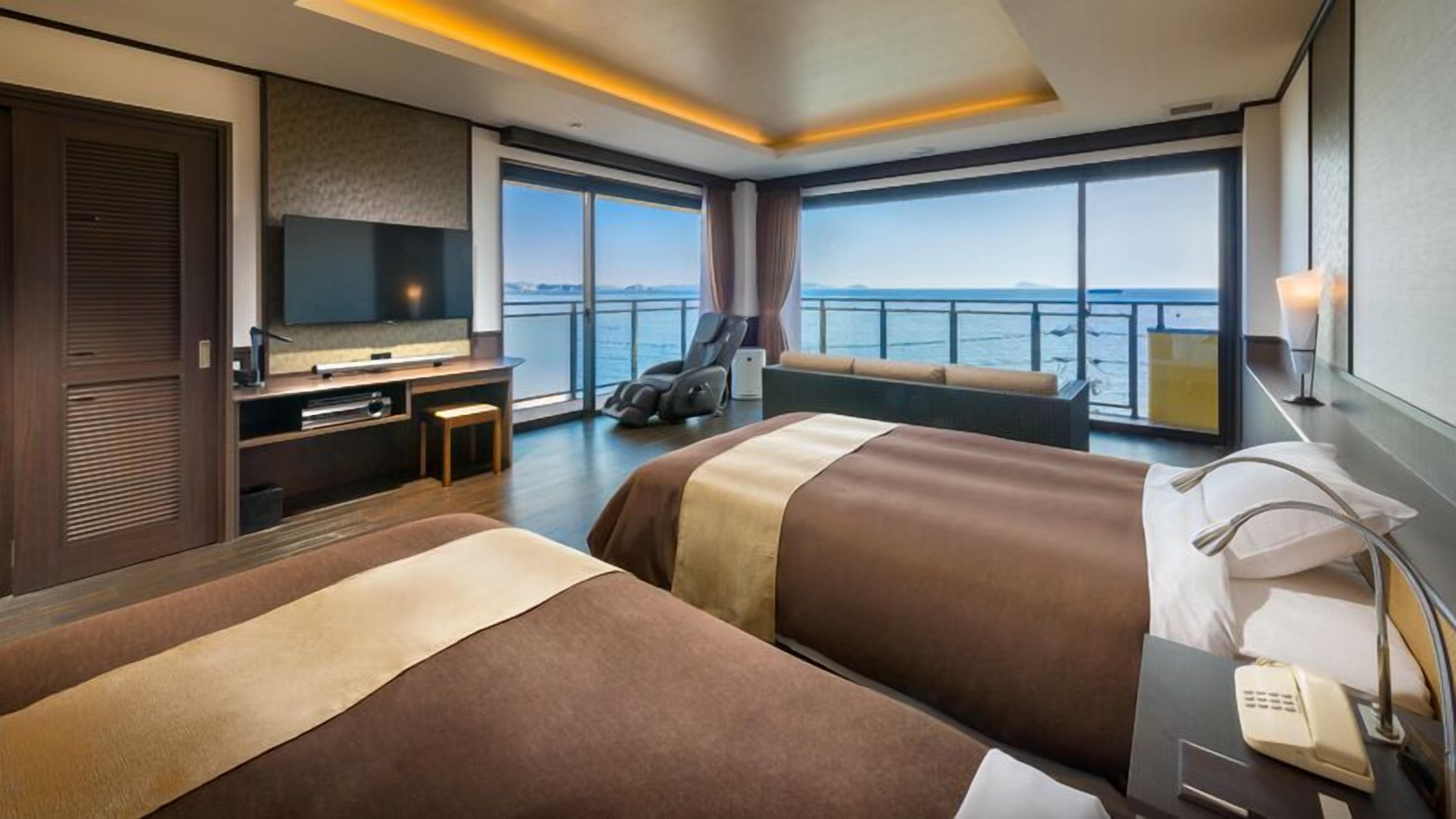 Ein Hotelzimmer mit zwei Betten, einem Fernseher, einem Massagesessel und Meerblick.