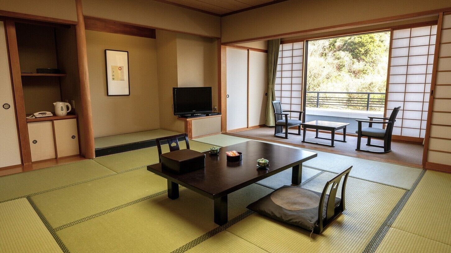 Ein traditioneller japanischer Raum mit Tatami-Böden, einem niedrigen Tisch mit Kissen, einem Fernseher und Schiebetüren, die auf einen Balkon führen.