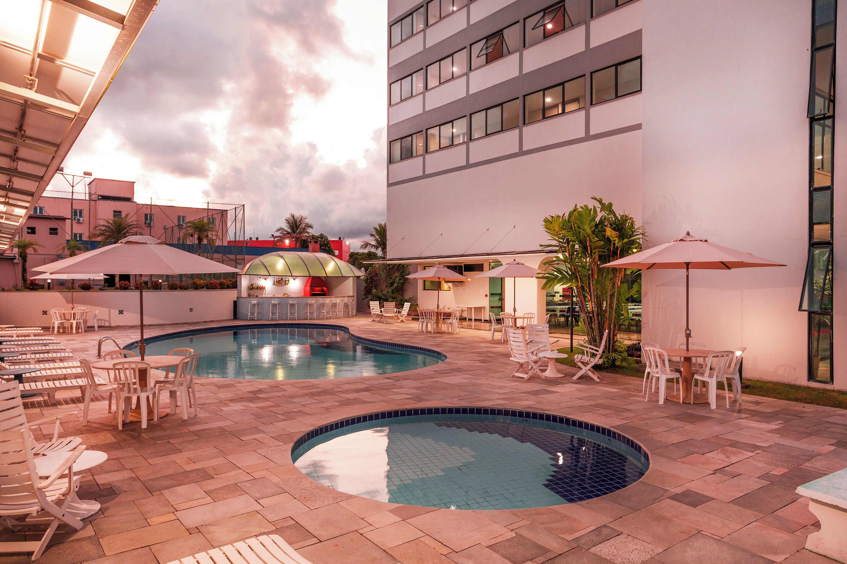 A pool area with lounge chairs and umbrellas, a building with multiple floors, and a clear sky.