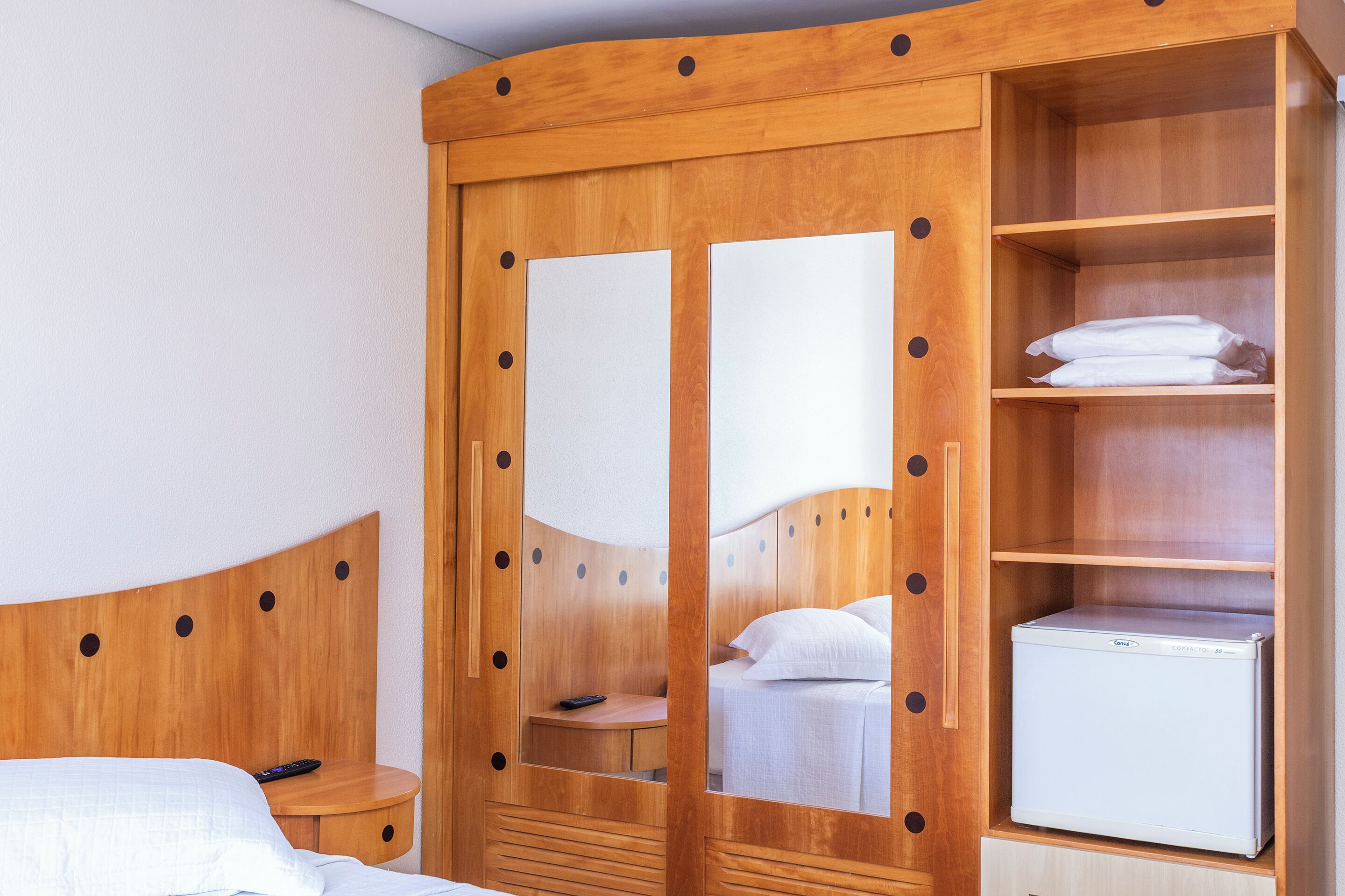 Um guarda-roupa de madeira com portas espelhadas, uma cama, uma mesa de cabeceira e um frigorífico branco.
