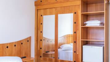 Um guarda-roupa de madeira com portas espelhadas, uma cama, uma mesa de cabeceira e um frigorífico branco.