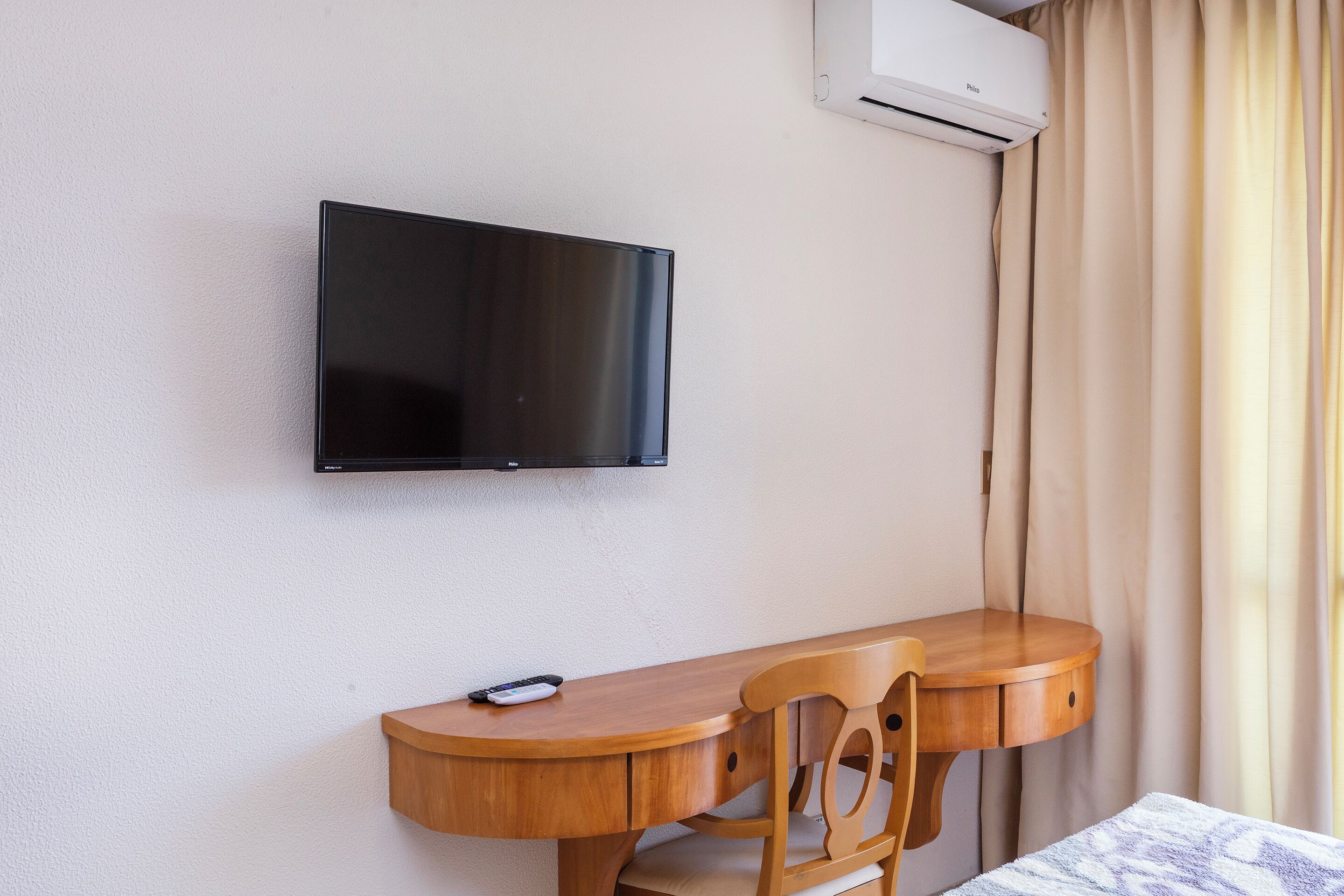 A room with a flat-screen TV mounted on the wall, a wooden desk with a chair, and an air conditioning unit.
