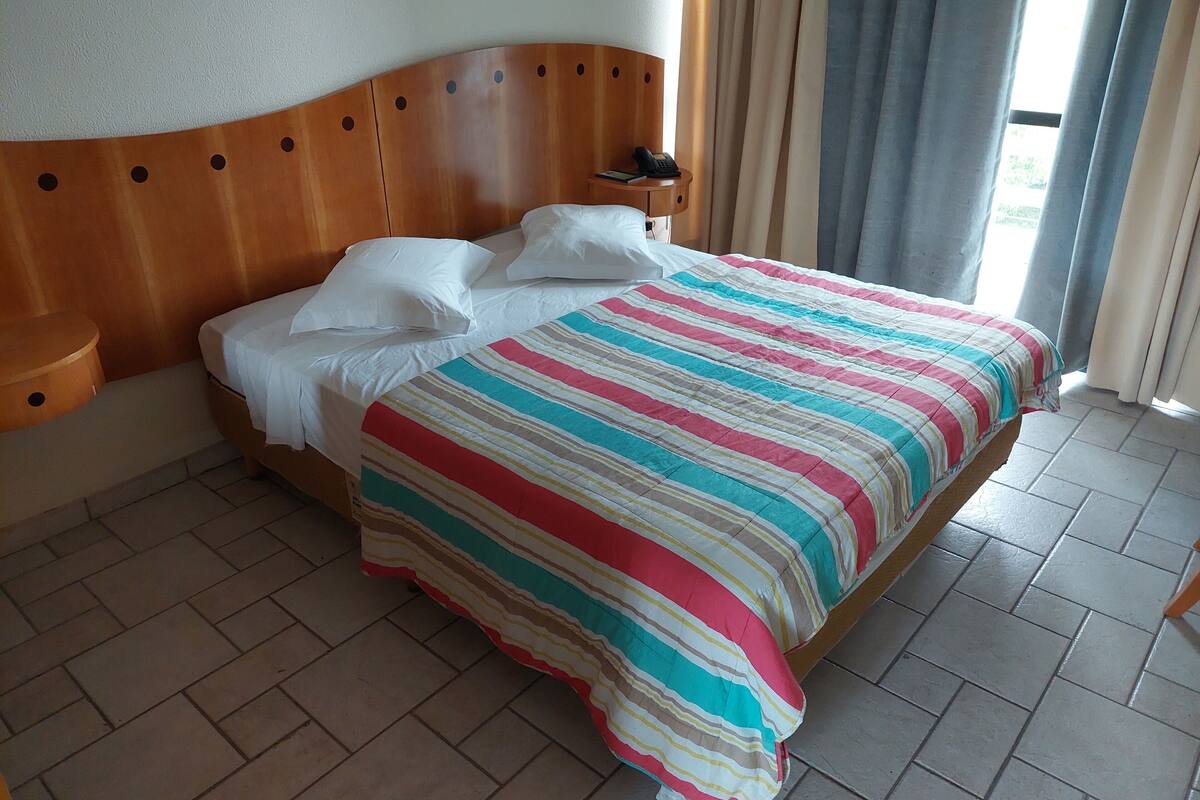 Une chambre d’hôtel avec une tête de lit en bois, une couvre-lit à rayures, des oreillers blancs, une table de chevet avec un téléphone et une fenêtre avec des rideaux.