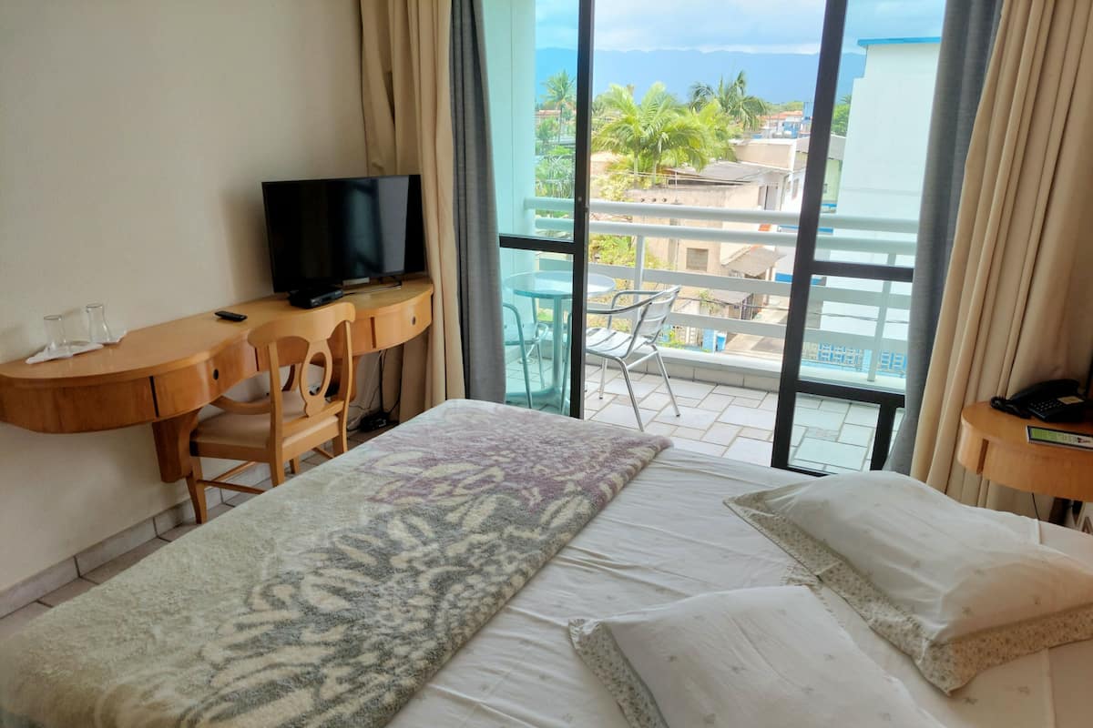 Une chambre d’hôtel comprenant un lit, un bureau avec une télévision, une chaise et un balcon offrant une vue sur les immeubles et les palmiers.