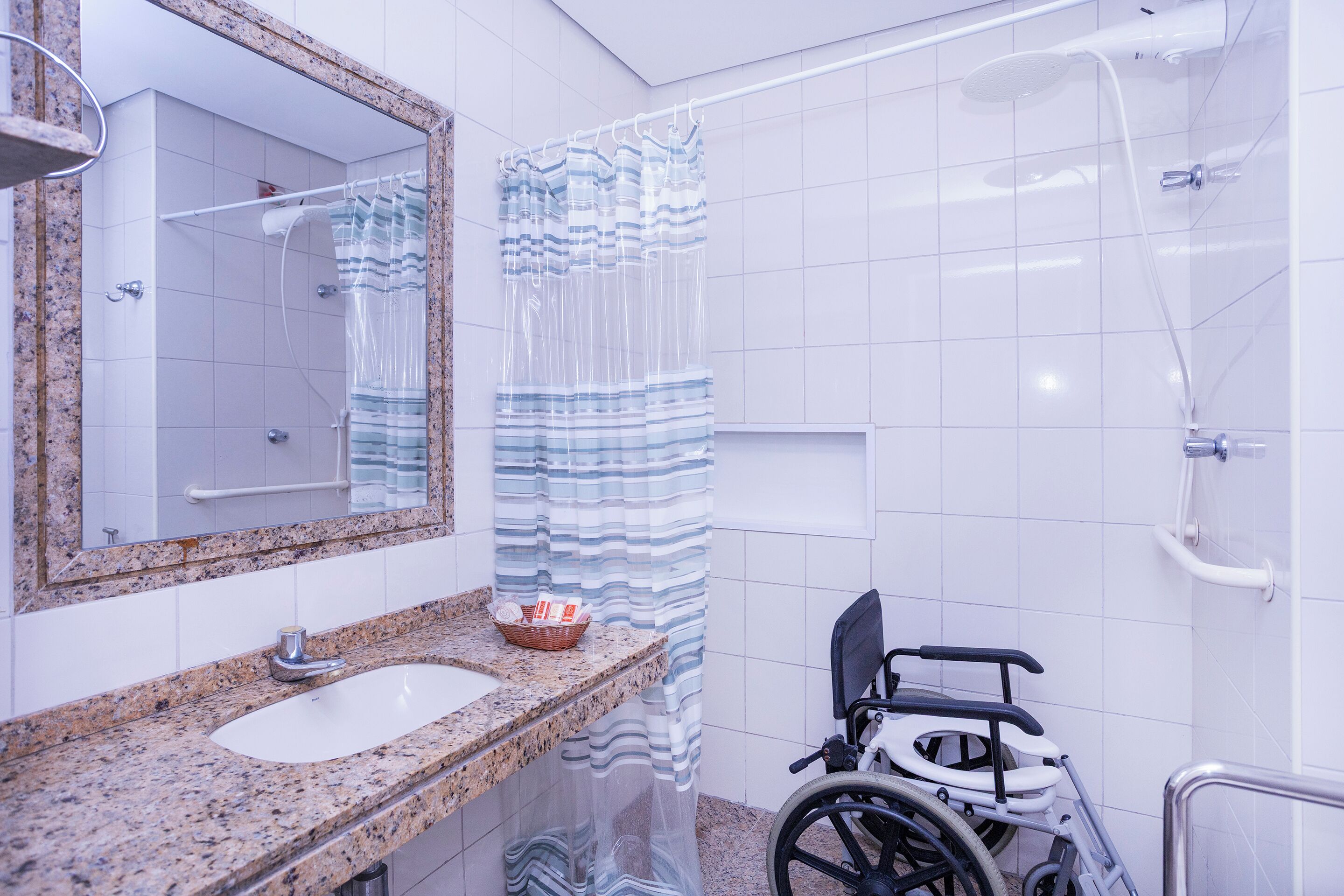 A bathroom with a granite countertop sink, a mirror, a shower with a transparent curtain, and a wheelchair.