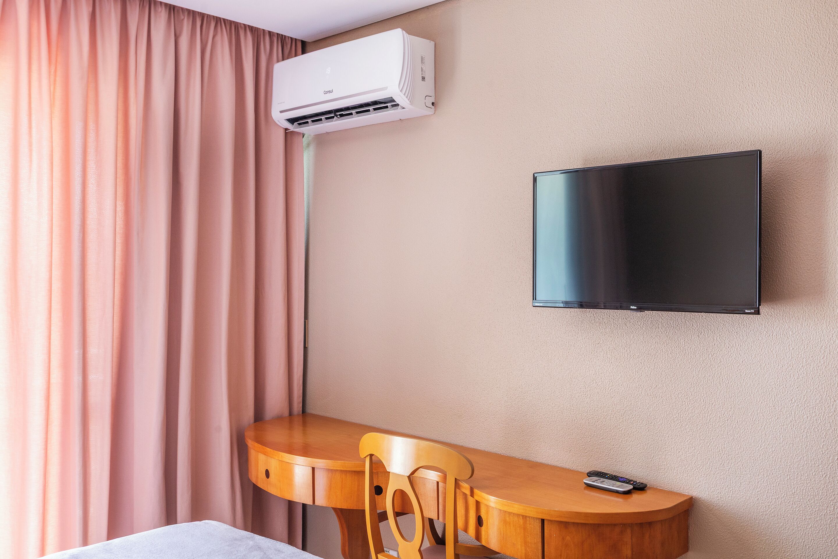 Habitación de hotel con televisor de pantalla plana montado en la pared, un escritorio de madera con una silla y un equipo de aire acondicionado sobre el escritorio.