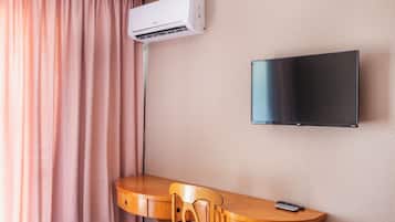 A hotel room with a flat-screen TV mounted on the wall, a wooden desk with a chair, and an air conditioning unit above the desk.