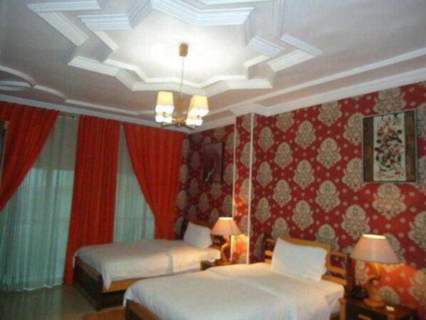 Una habitación de hotel con dos camas, un candelabro, cortinas rojas, un papel tapiz estampado y un cuadro en la pared.