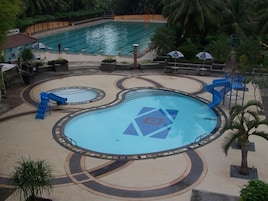 Un área de piscina con varias piscinas, incluyendo una piscina central con diseño geométrico, rodeada de zonas de descanso y palmeras.