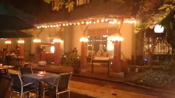 An outdoor dining area with tables and chairs, a covered seating area with string lights, and a building entrance.