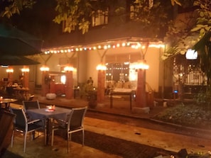 Ein Außenbereich zum Essen mit Tischen und Stühlen, eine überdachte Sitzgelegenheit mit Lichterketten und ein Gebäudeeingang.