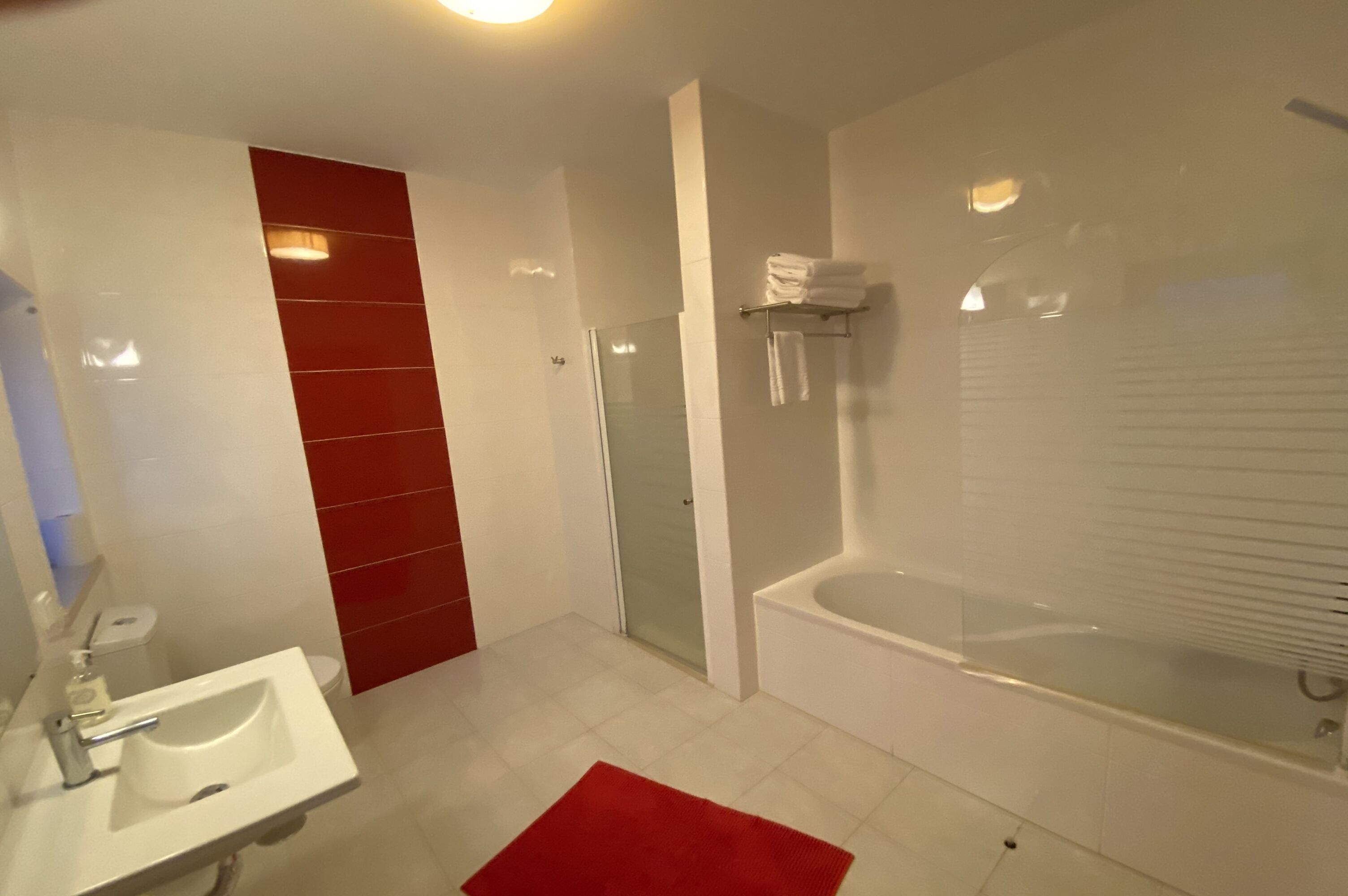 Un baño moderno con un lavamanos blanco, pared con azulejos rojos, cabina de ducha de vidrio y una bañera blanca.