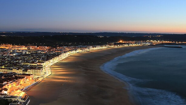 An aerial view of a coastal city with a long beachfront, illuminated buildings, and a clear sky. - Villas Mare Residence (Nazaré)
