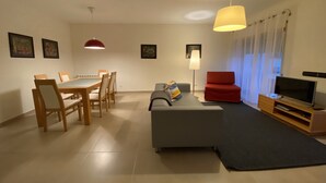 A modern living room with a dining area, a grey sofa, a red chair, a TV, and a lamp. - Villas Mare Residence (Nazaré)