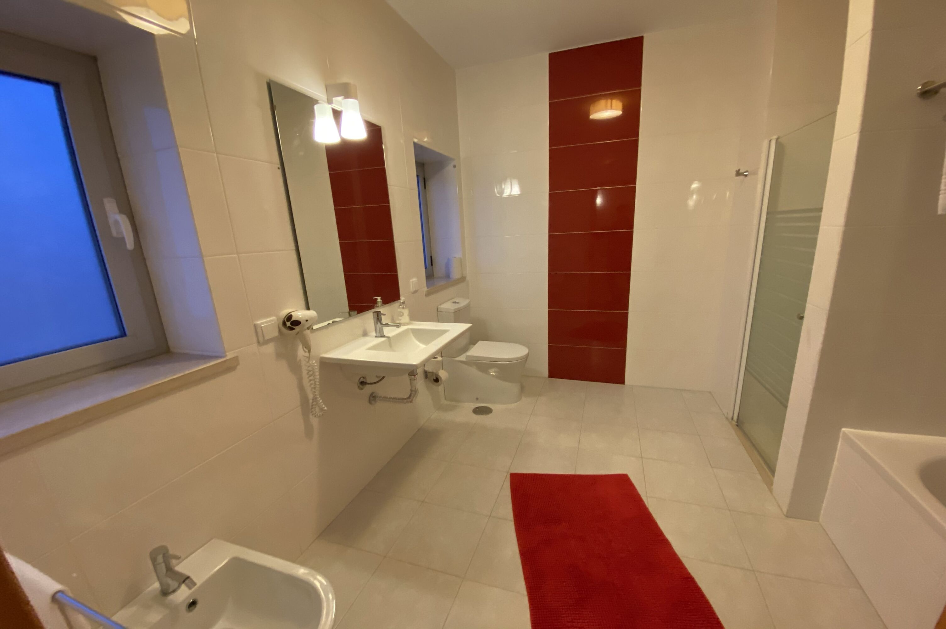 Un baño moderno con lavamanos, inodoro y bañera blancos, una pared de acento roja y una cabina de ducha con paredes de vidrio.