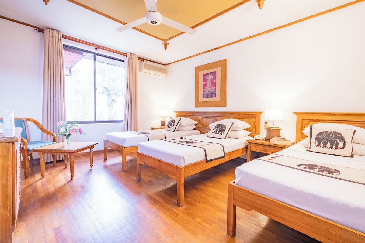 A hotel room with a wooden floor, two beds with elephant-themed pillows, a desk with a chair, a window with curtains, and a ceiling fan.