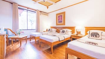 A hotel room with a wooden floor, two beds with elephant-themed pillows, a desk with a chair, a window with curtains, and a ceiling fan.