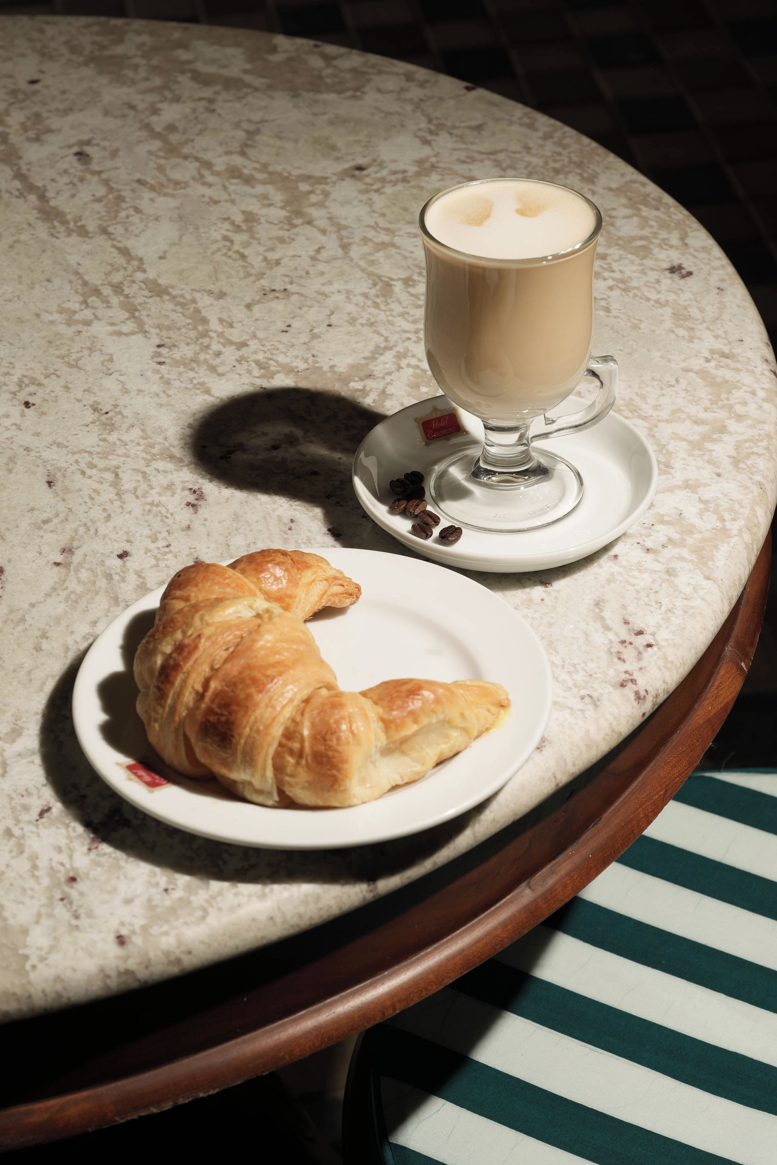 A croissant on a plate, a cup of coffee, and a small bowl of coffee beans on a marble table.
