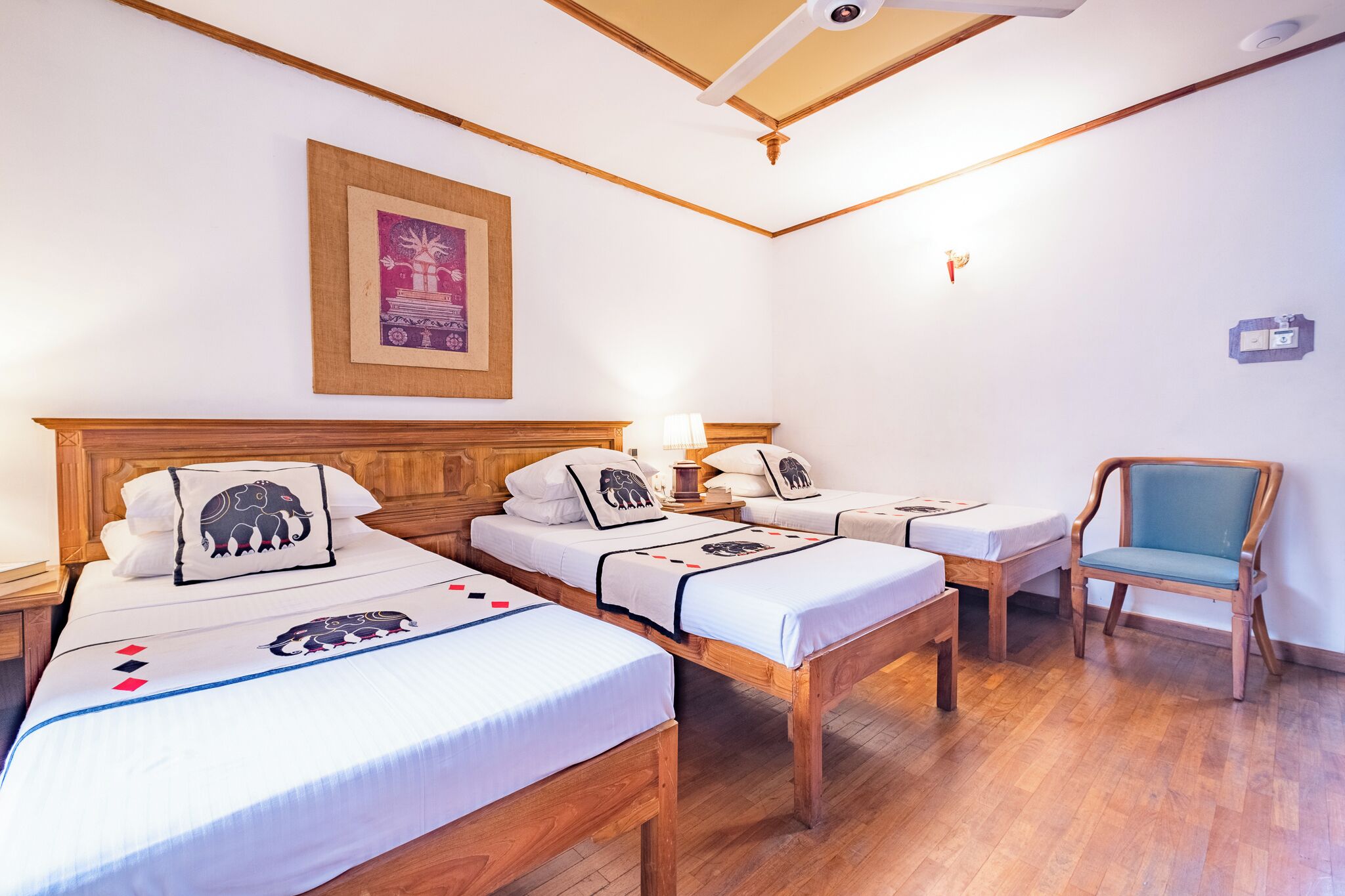Habitación de hotel con tres camas, una silla de madera, un cuadro enmarcado en la pared y una lámpara en el techo.