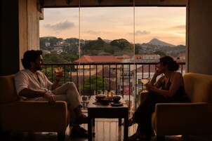 Dos personas sentadas en un balcón con vista a la ciudad, una mesa con comida y bebidas, y un atardecer al fondo.