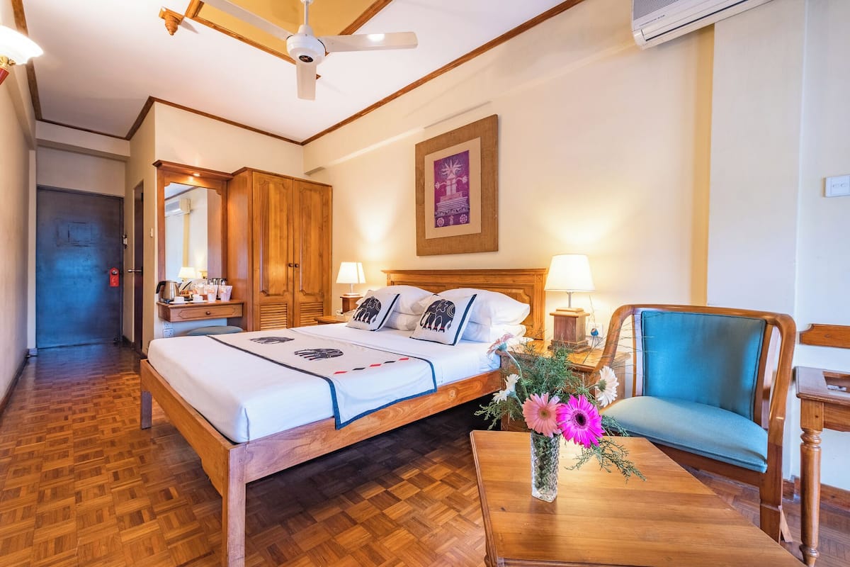 A hotel room with a wooden bed, two bedside lamps, a wooden chair, a wooden table, and a ceiling fan.