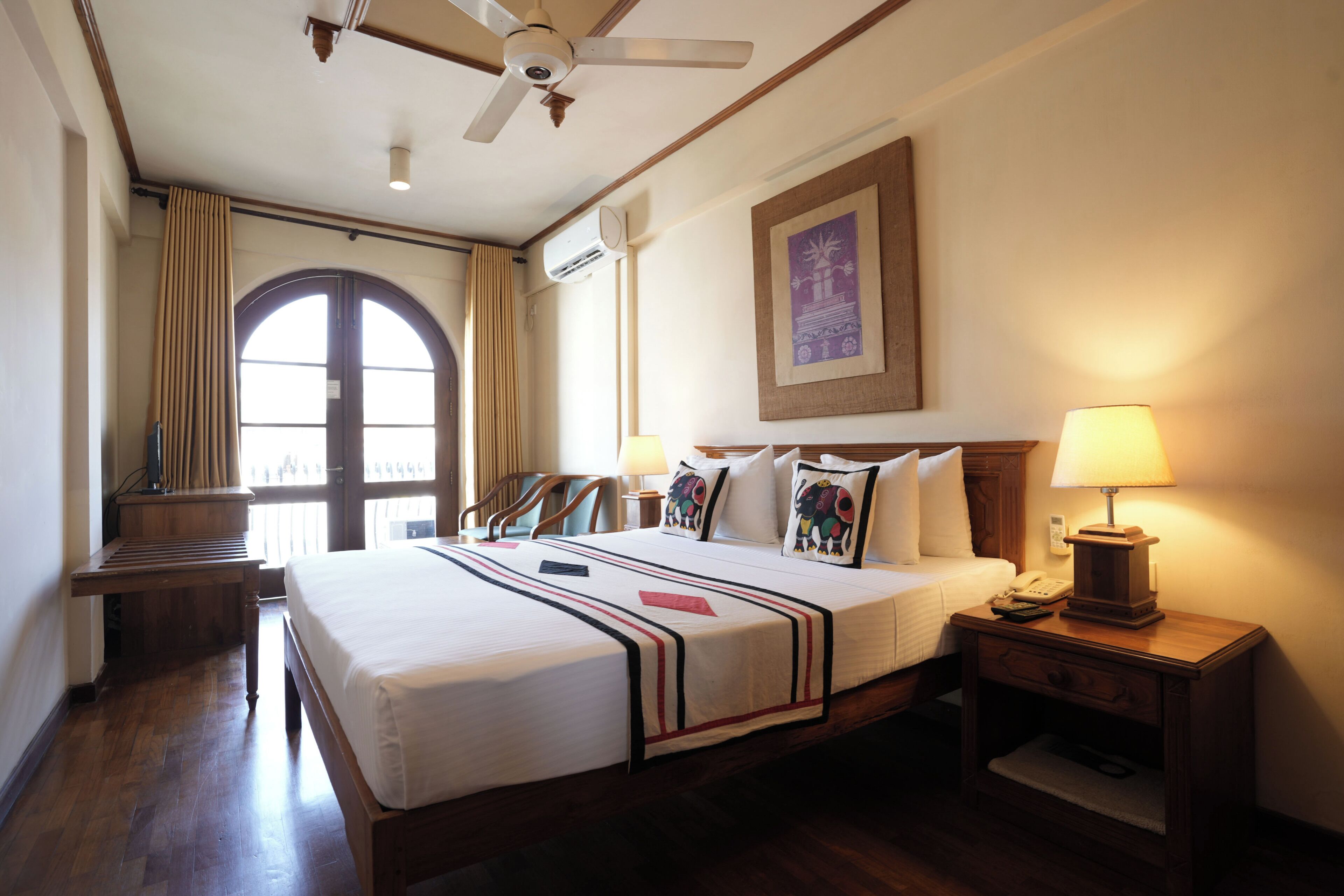 A hotel room with a bed, a nightstand, a lamp, a ceiling fan, an air conditioning unit, a window with curtains, and a framed picture on the wall.