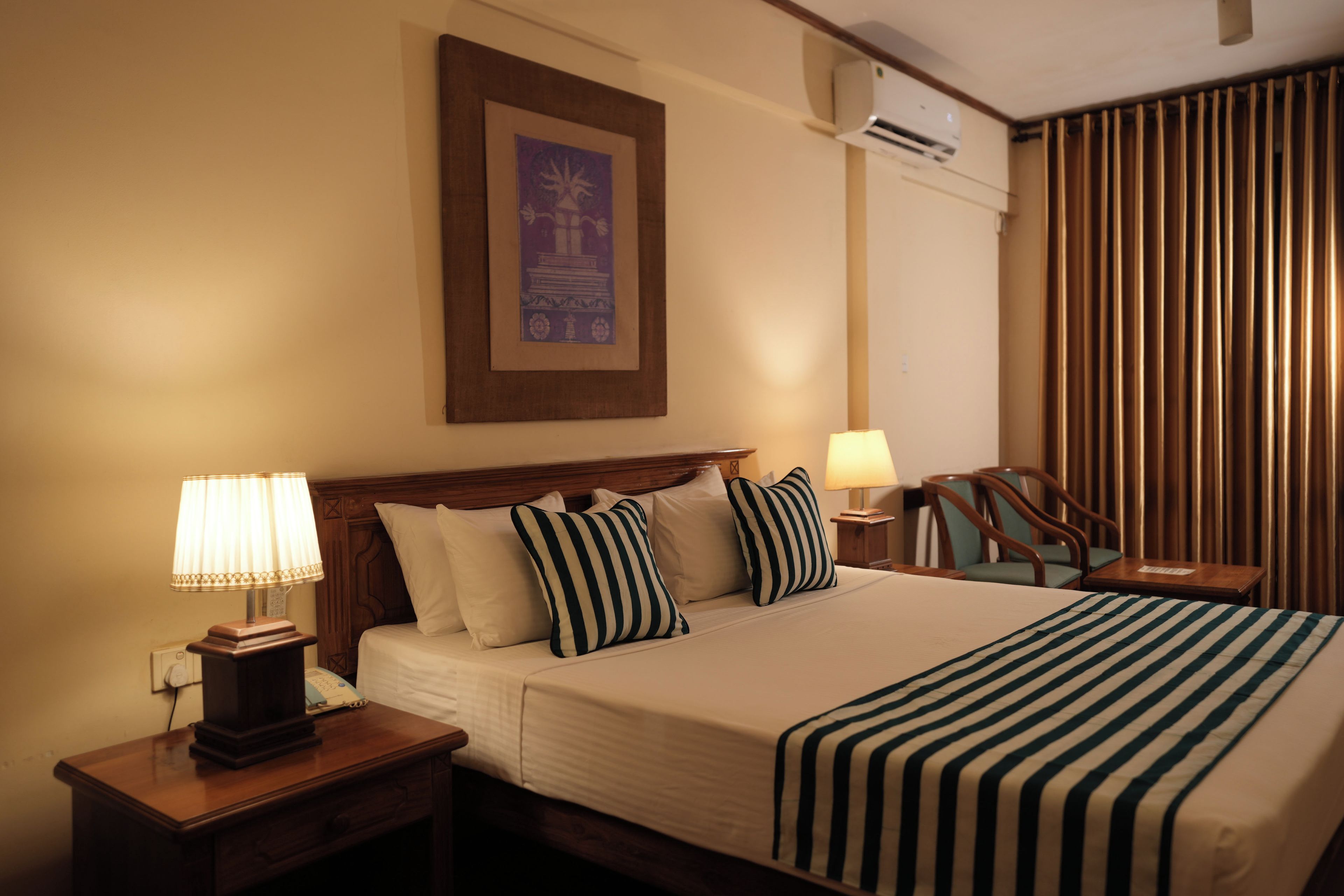 Une chambre d’hôtel avec un lit, deux tables de chevet avec des lampes, une tête de lit en bois, un tableau encadré au mur, un appareil de climatisation et une fenêtre avec des rideaux.
