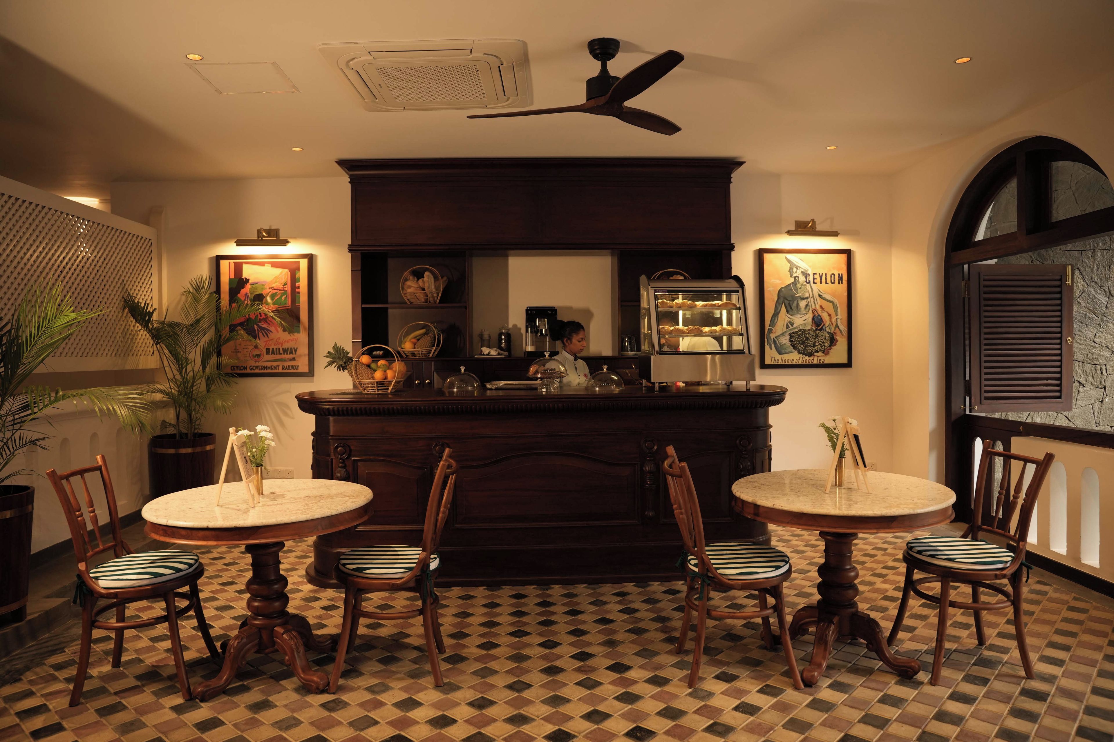 A restaurant interior with a bar, two round tables with chairs, a ceiling fan, and wall-mounted artwork.