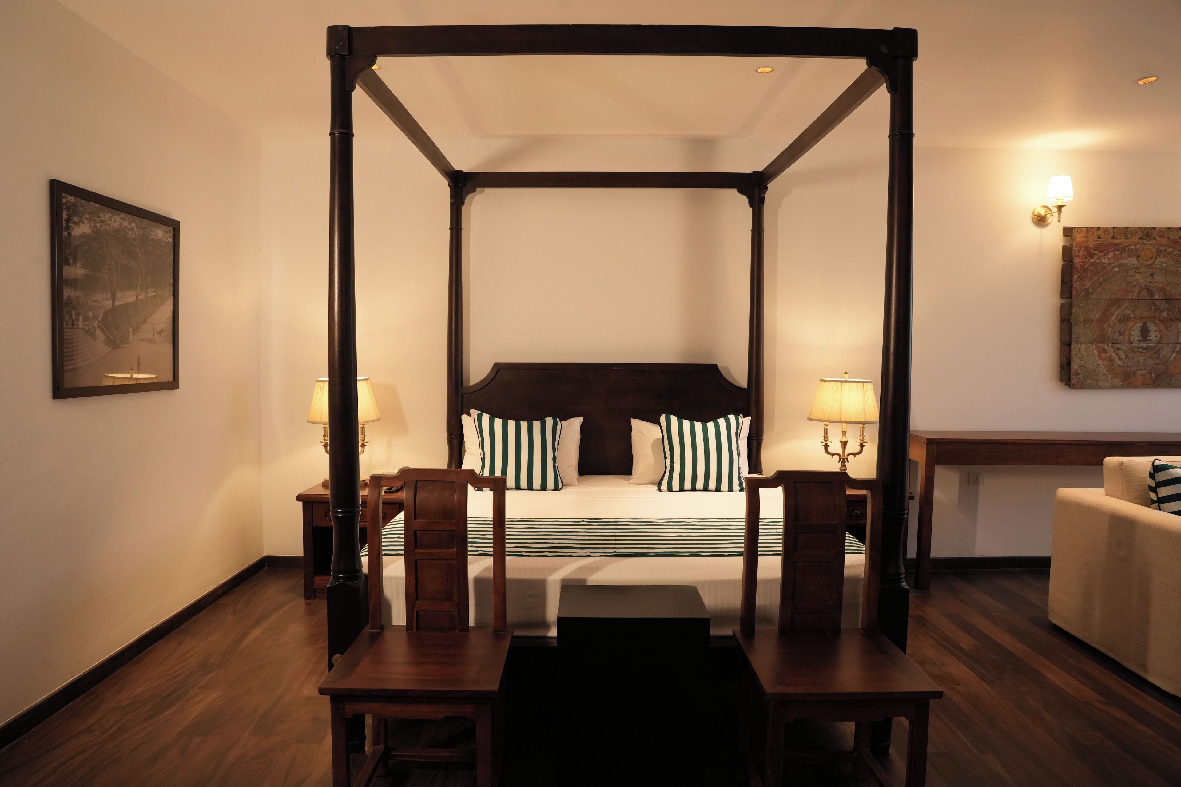 Un lit à baldaquin avec une literie à rayures, deux tables de chevet avec des lampes, une chaise en bois, une petite table et un tableau accroché au mur.