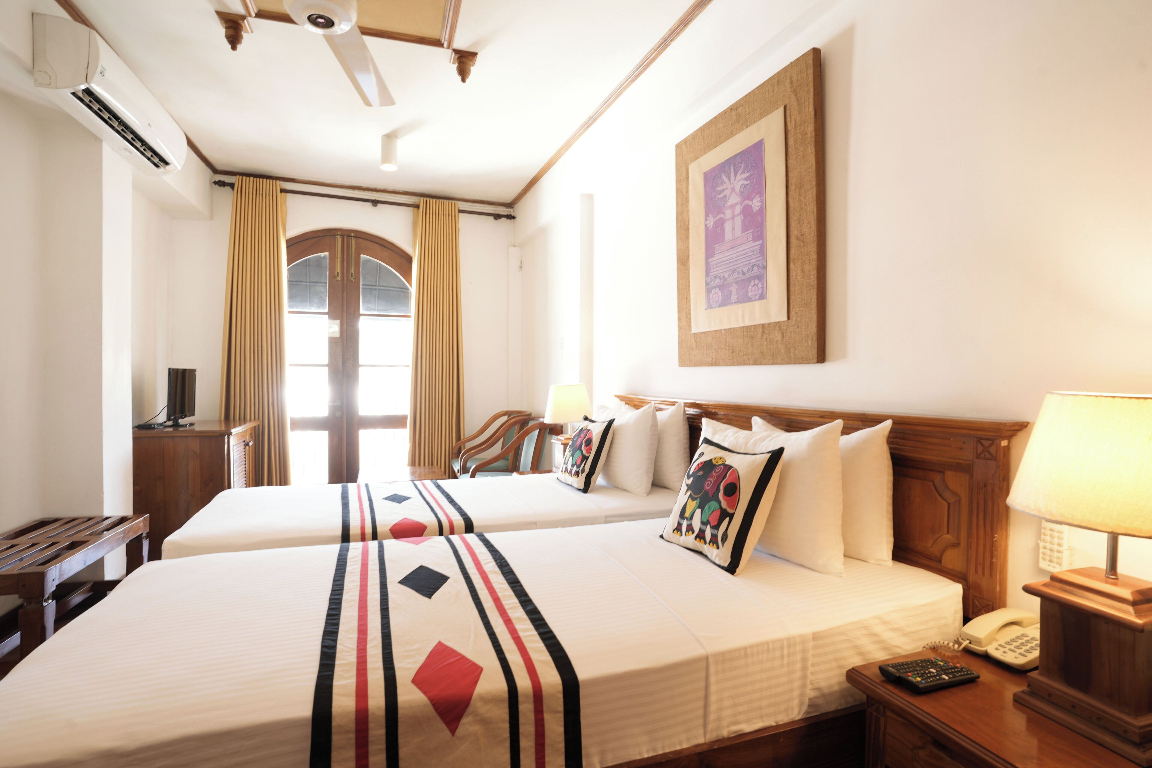 A hotel room with two beds, a wooden headboard, a desk with a lamp, a telephone, and a window with curtains.