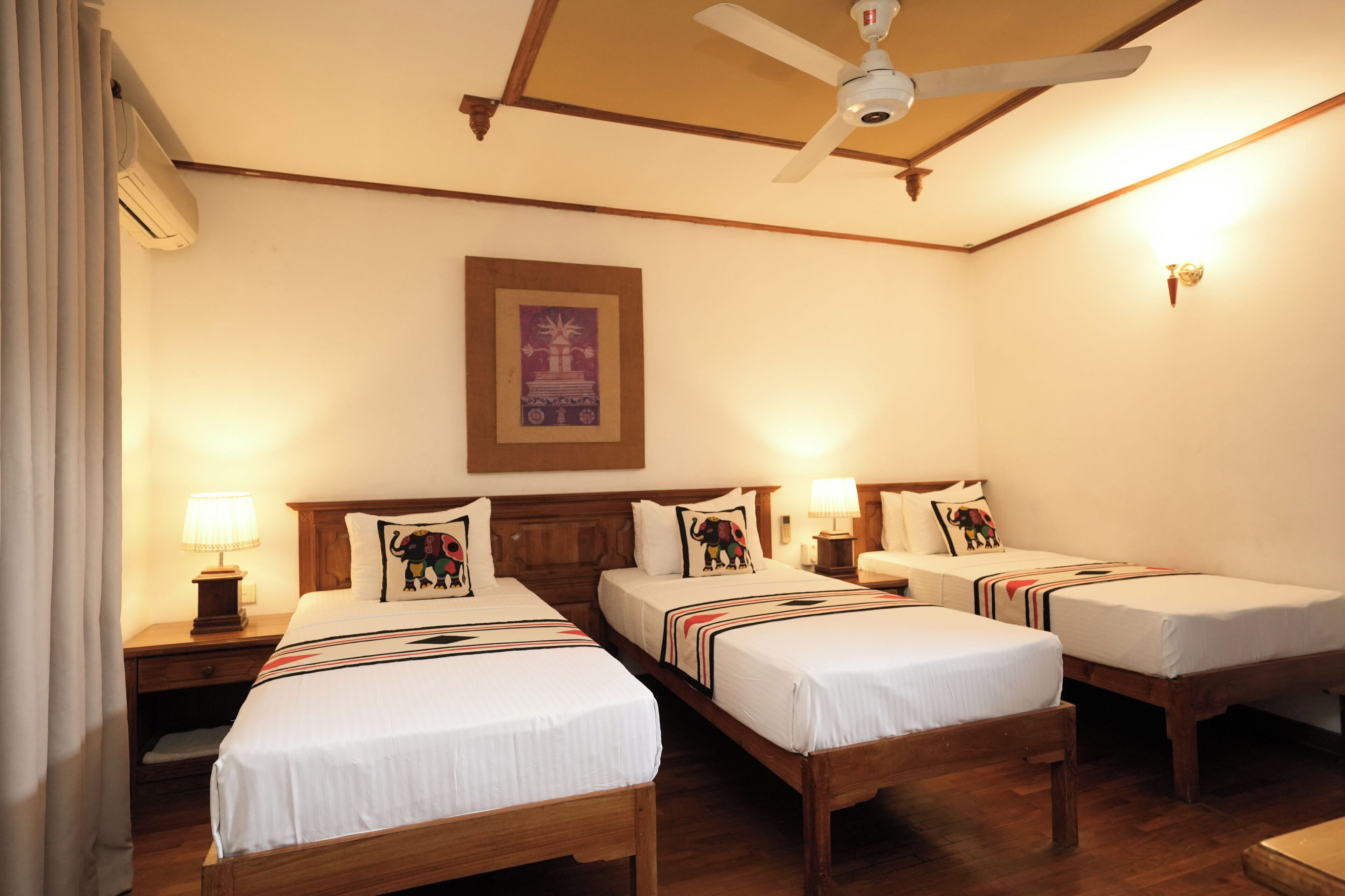 A hotel room with three wooden beds, a ceiling fan, and a framed picture on the wall.